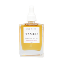 Wild Rising Tamed Conditioning Hair Oil in a clear square glass bottle with a white dropper cap, filled with golden-yellow oil, on a white background.