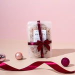 A packaged bath salt product with a label reading 'Awaken' tied with a red ribbon, placed on a surface with a pink background and decorative elements around it.
