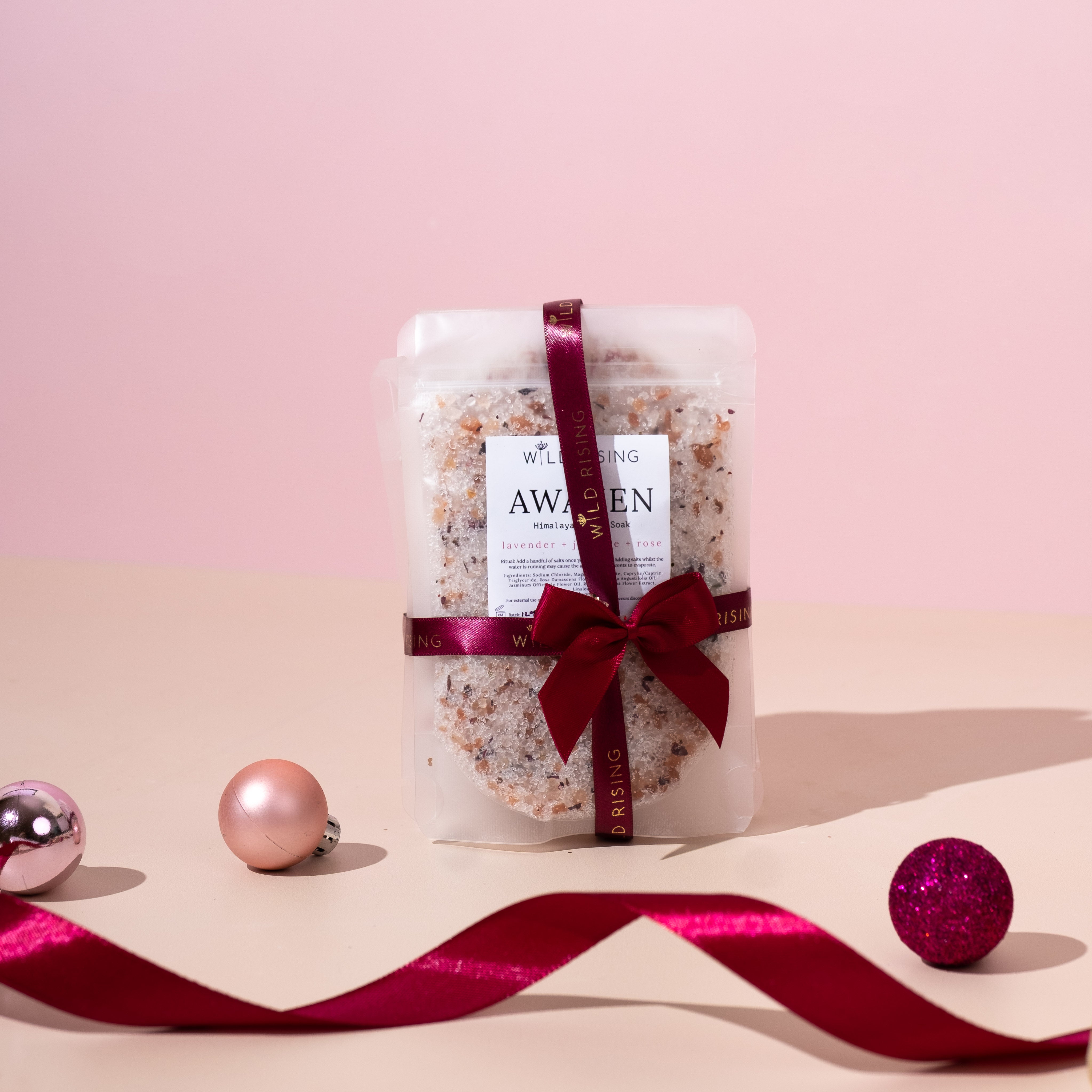 A packaged bath salt product with a label reading 'Awaken' tied with a red ribbon, placed on a surface with a pink background and decorative elements around it.