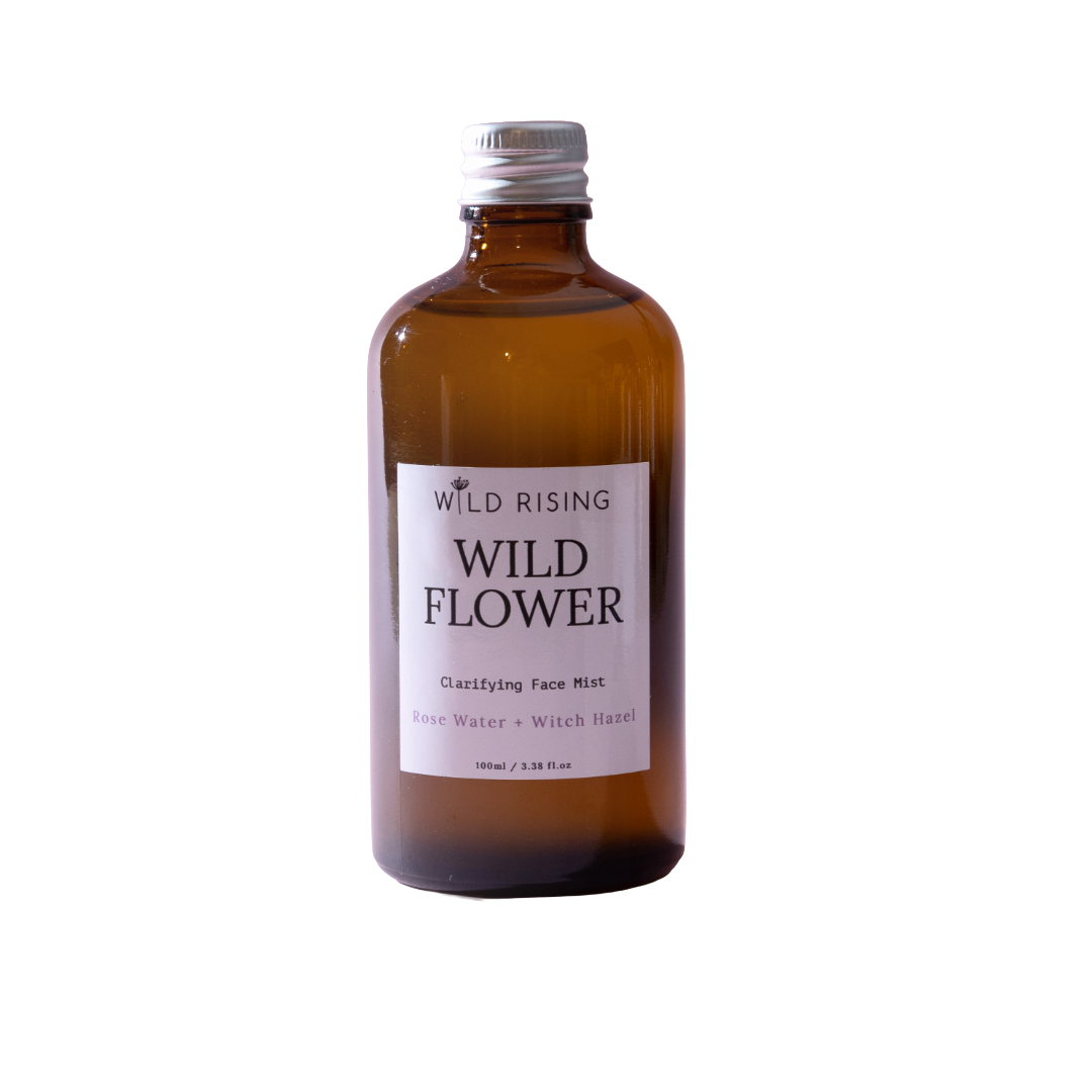 An amber glass bottle of Wild Flower Clarifying Face Mist by The Wild Rising, featuring a white label and a silver metal cap, set against a plain white background.