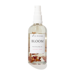 A clear spray bottle of Wild Rising Bloom Nourishing Body Oil with a white pump dispenser. The transparent bottle shows a light beige oil inside with dried coconut and rose petals suspended at the bottom, set against a clean white background with a soft shadow.
