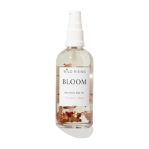 A clear spray bottle of Wild Rising Bloom Nourishing Body Oil with a white pump dispenser. The transparent bottle shows a light beige oil inside with dried coconut and rose petals suspended at the bottom, set against a clean white background with a soft shadow.