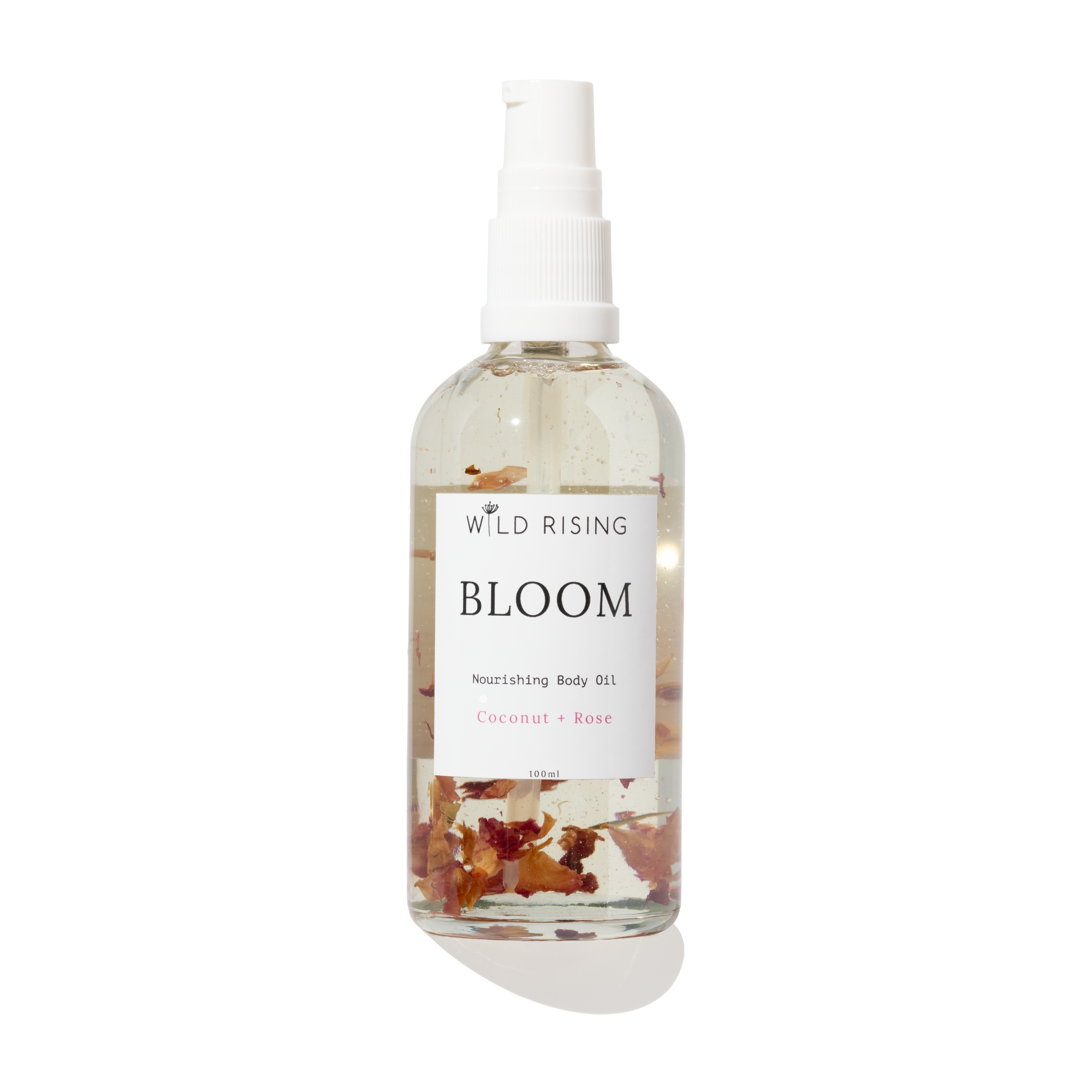 A clear spray bottle of Wild Rising Bloom Nourishing Body Oil with a white pump dispenser. The transparent bottle shows a light beige oil inside with dried coconut and rose petals suspended at the bottom, set against a clean white background with a soft shadow.
