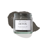 A glass jar of Wild Rising Detox Activated Charcoal Mask, filled with dark green French clay powder and topped with a small pile of the same charcoal mask, set against a clean white background.
