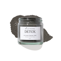 A glass jar of Wild Rising Detox Activated Charcoal Mask, filled with dark green French clay powder and topped with a small pile of the same charcoal mask, set against a clean white background.
