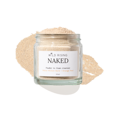 A clear glass jar labeled 'WILD RISING NAKED' containing a light beige powder, set against a white background with a matching beige powder smear behind it.