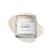 A clear glass jar labeled 'WILD RISING PURIFY Botanical Face Mask' filled with a light beige powder, set against a white background with a matching powder spill behind it.