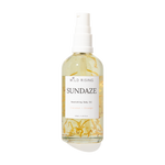 A 100ml spray bottle of Wild Rising Sundaze Coconut + Orange nourishing body oil, filled with pale yellow oil and suspended golden botanical pieces, set against a plain white background.