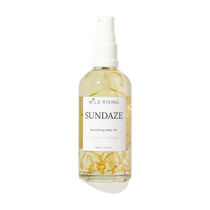 A 100ml spray bottle of Wild Rising Sundaze Coconut + Orange nourishing body oil, filled with pale yellow oil and suspended golden botanical pieces, set against a plain white background.