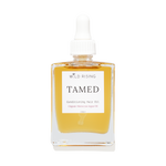 Wild Rising Tamed Conditioning Hair Oil in a clear square glass bottle with a white dropper cap, filled with golden-yellow oil, on a white background.