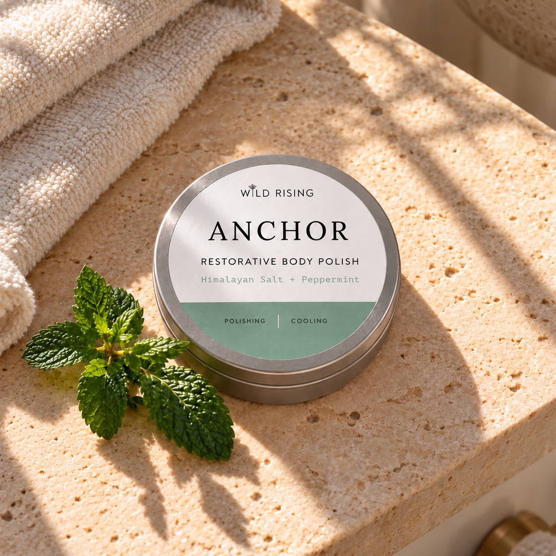 A round tin of WILD RISING ANCHOR Restorative Body Polish sits on a beige stone surface next to a green mint sprig and a folded cream towel, with warm natural sunlight casting shadows.