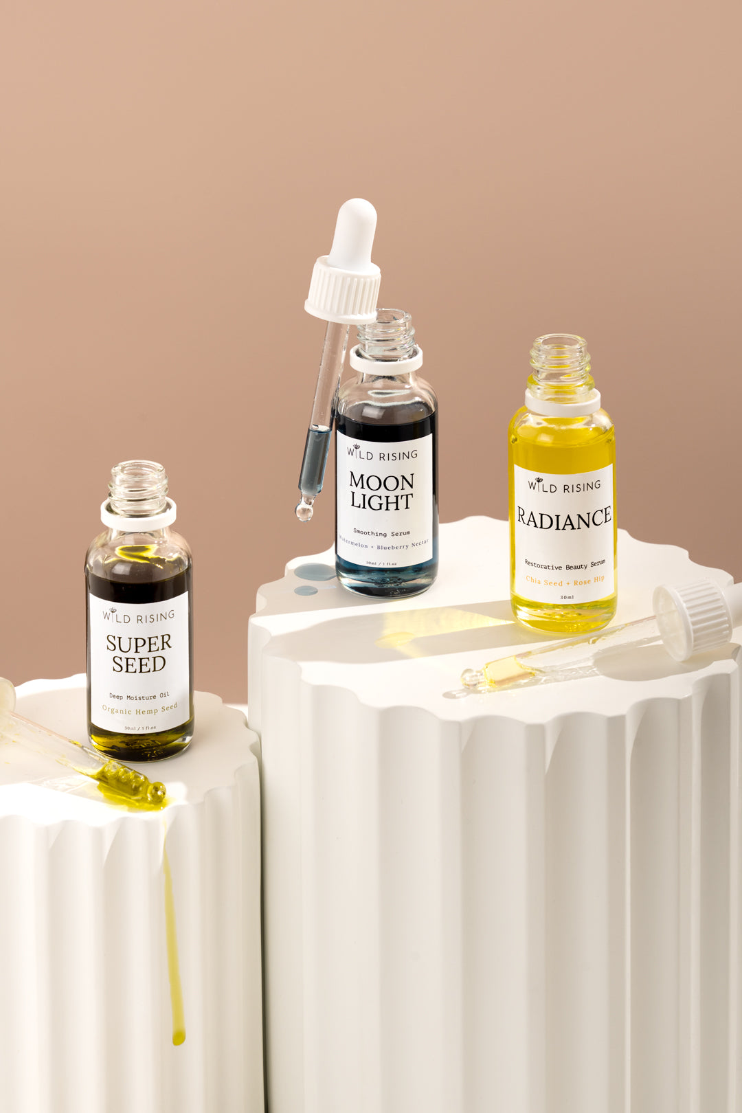 Three skincare serum bottles from Wild Renskin displayed on white pedestal stands against a warm beige background. The bottles contain dark brown, black, and yellow liquids and are labeled 'Super Seed', 'Moon Light', and 'Radiance'.