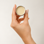 A hand with manicured nails holds an open silver tin of cream-colored balm or lip balm against a plain light beige background.