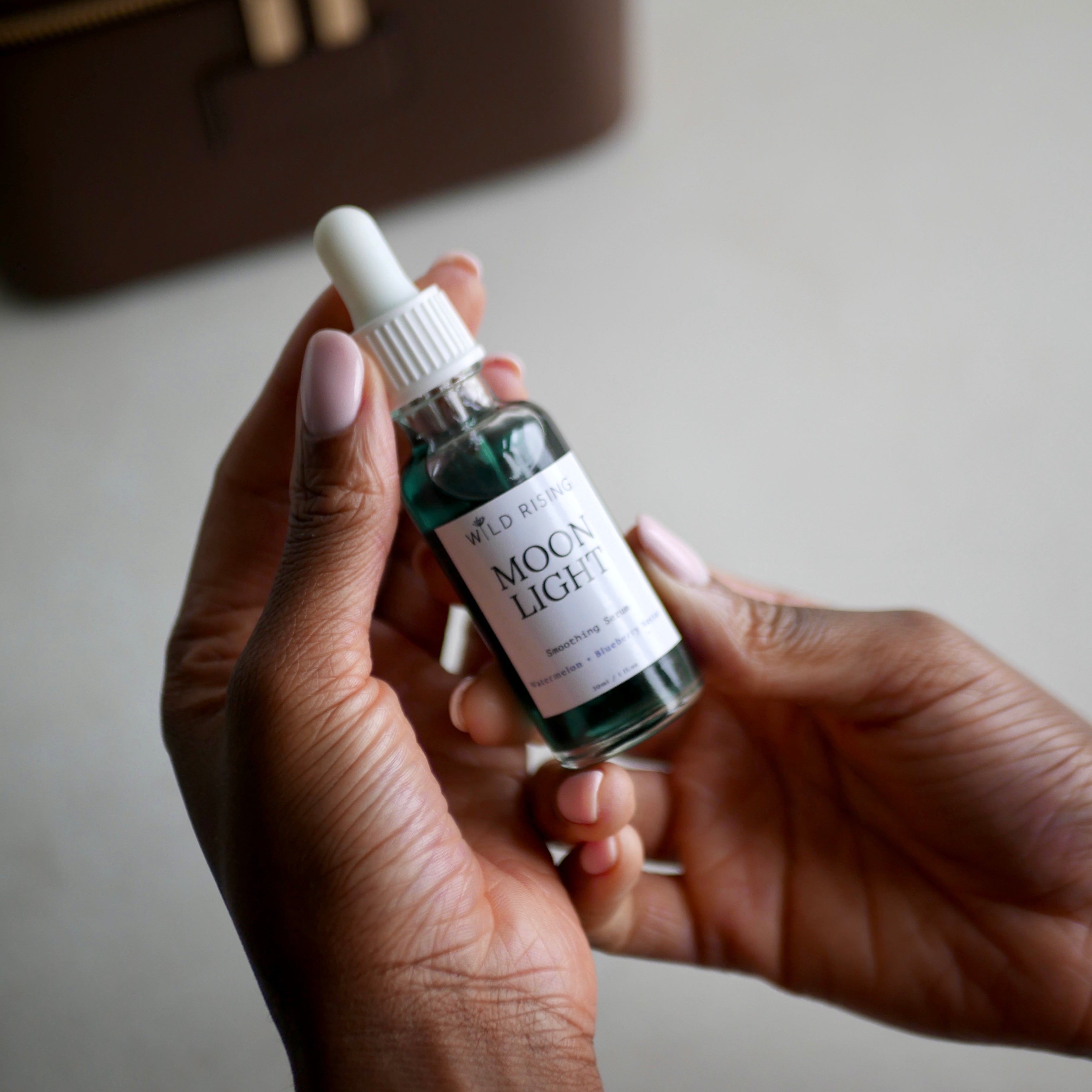 A person's hands holding a small glass dropper bottle labeled 'MOON LIGHT' against a soft beige background with a brown case blurred in the distance.