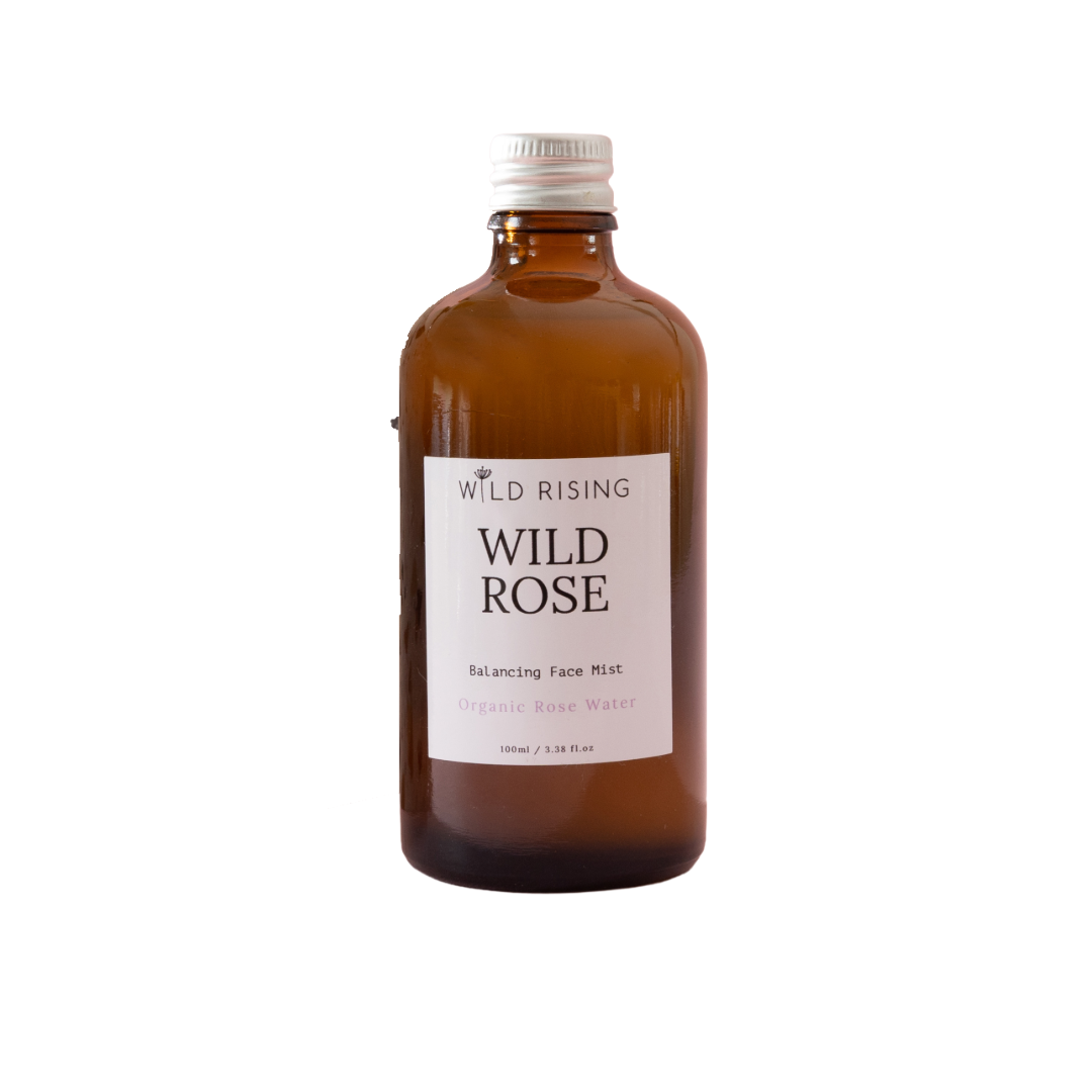 An amber glass bottle of Wild Rising Wild Rose Balancing Face Mist with a white label and silver cap, isolated on a white background.