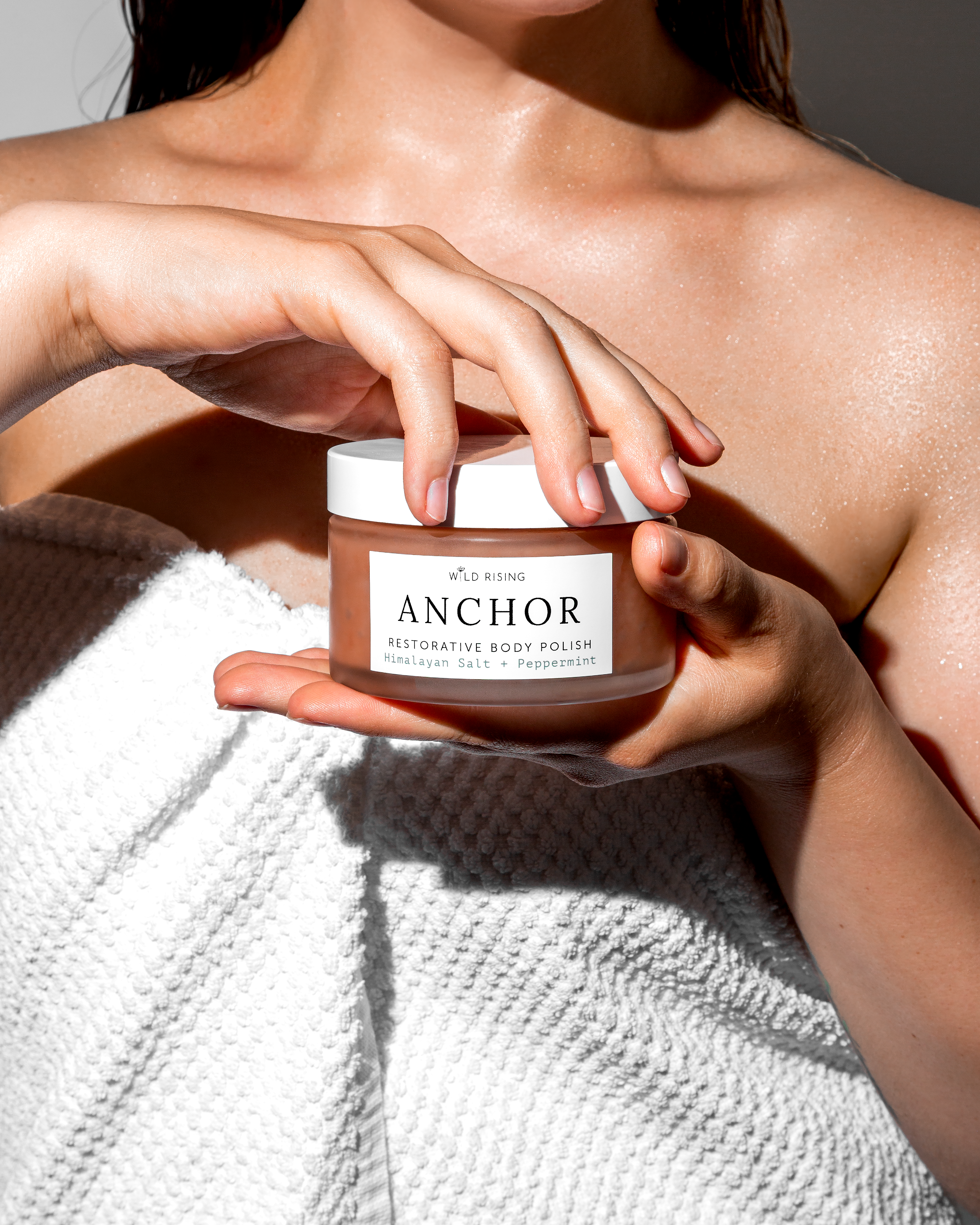 A close-up of a person wrapped in a white textured towel, holding a jar of ANCHOR Alternative Body Polish. The product is a clear jar showing a warm brownish-beige body polish inside, with a white lid and a white label featuring black text.