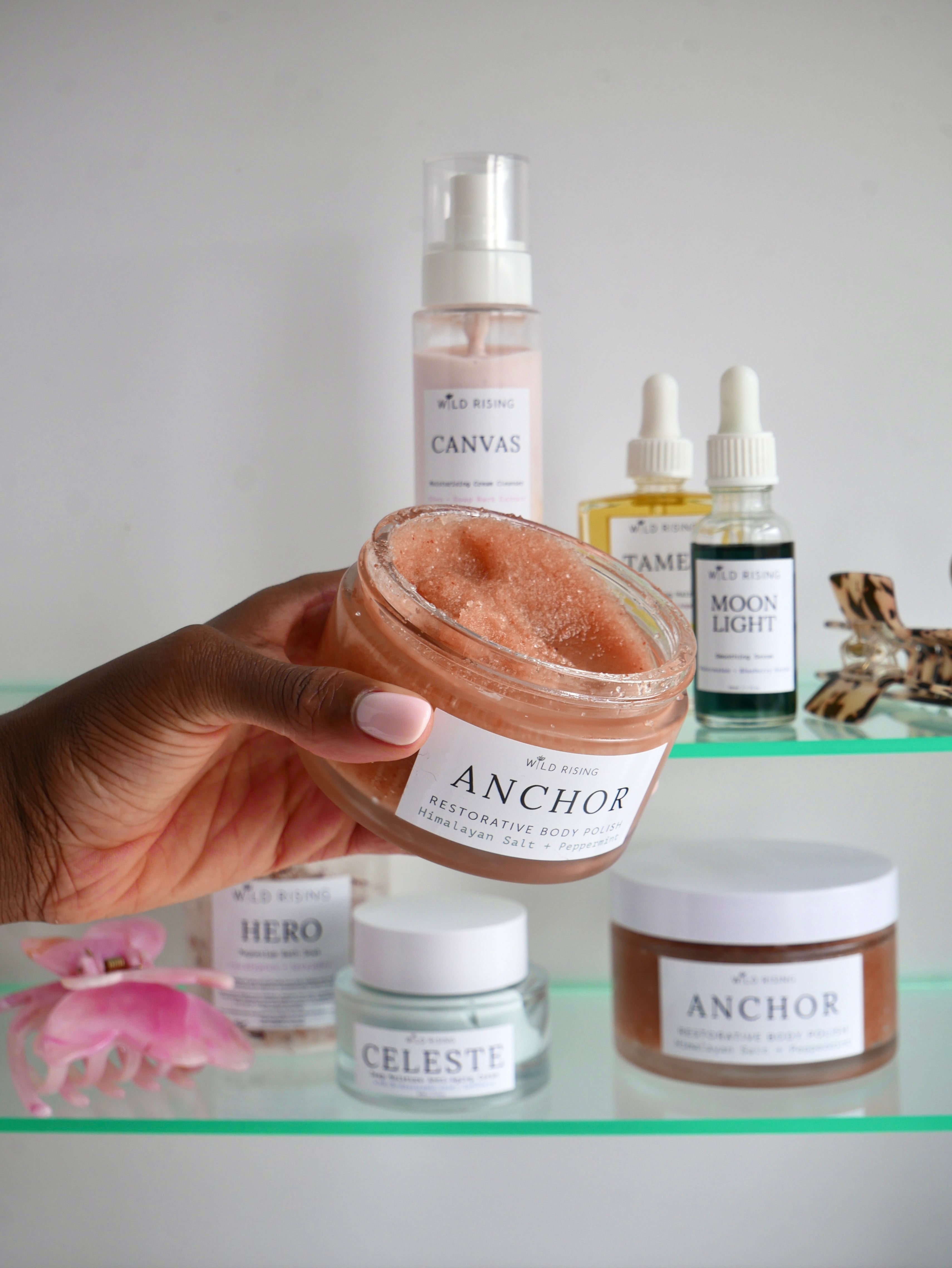 Hand holding a jar of 'Anchor' scrub with skincare products on a shelf in the background.