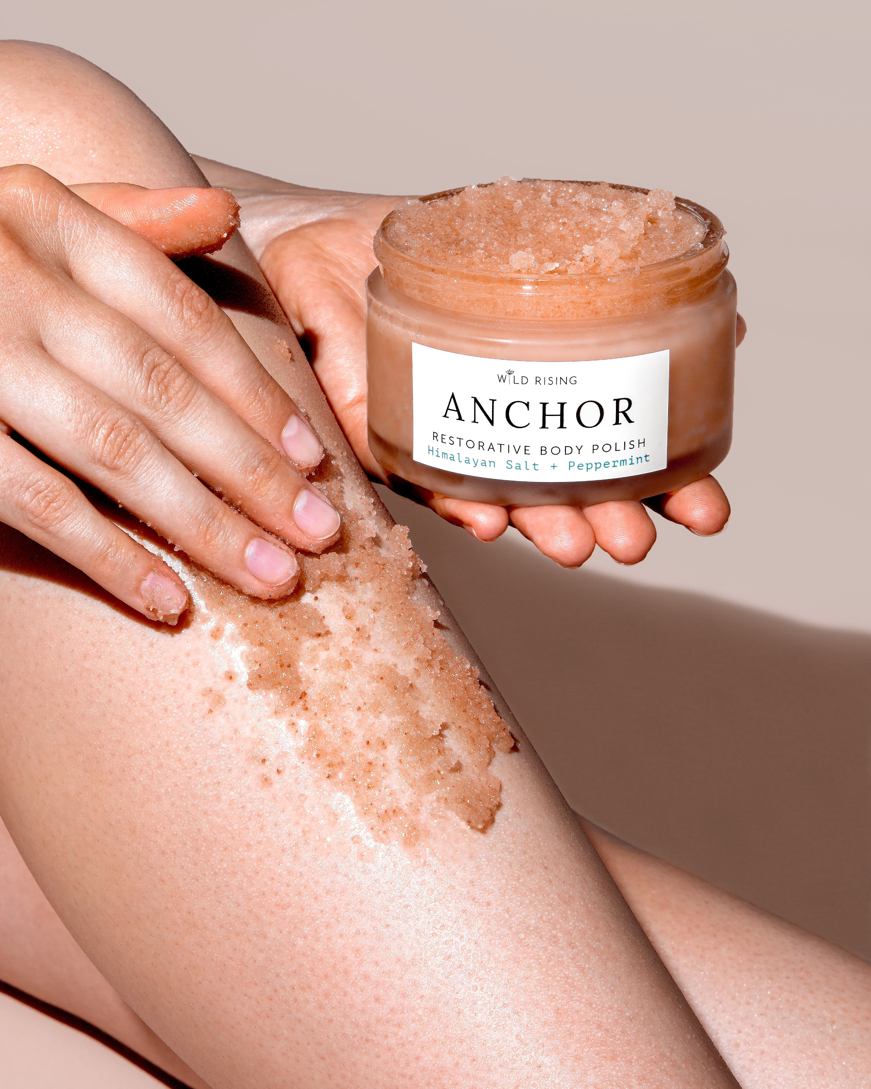 A person applies a brown exfoliating body polish from a clear jar labeled 'Wild Rising Anchor Restorative Body Polish' onto their leg, set against a solid beige background.