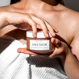 A person holding a jar of Wild Rising Anchor Restorative Body Polish with a white towel wrapped around their torso, under bright natural sunlight.