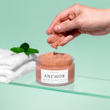 A hand holds a clump of pink exfoliating body polish above an open jar labeled Wild Being Anchor. The jar sits on a glass shelf beside folded white towels and mint leaves against a pale mint-green background.